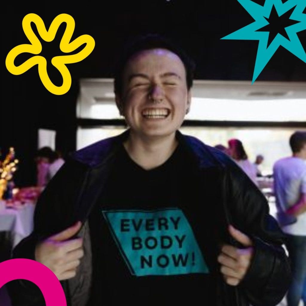 A person with short dark hair wearing an everybpdy now t-shirt smiles wide for the camera